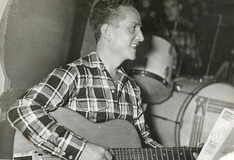 À la guitare, chemise à carreaux, devant la batterie de l'orchestre.